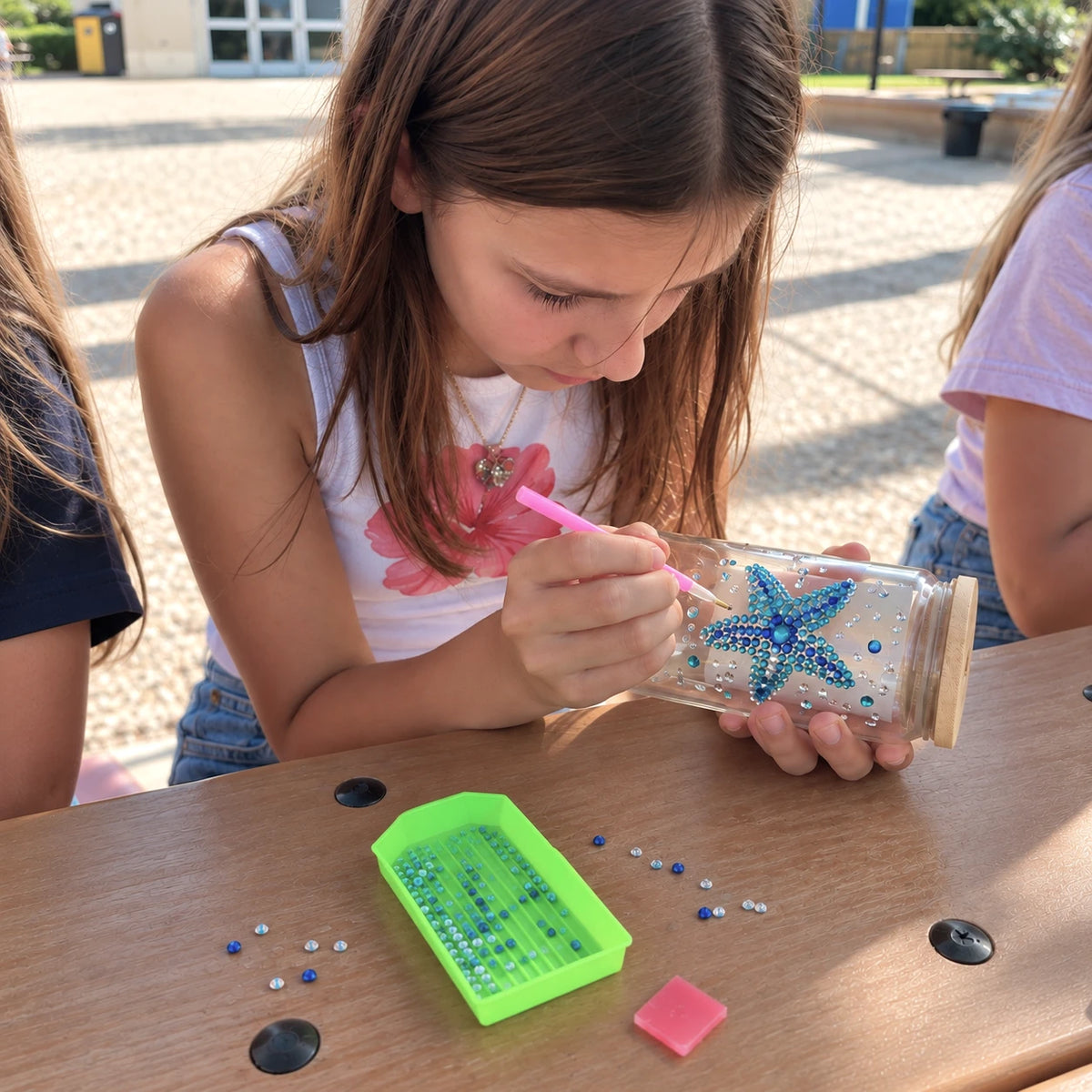 A young girl with long brown hair is sitting at a wooden table outdoors, carefully placing blue and silver rhinestones onto a clear jar at a mobile art workshop in Los Angeles.