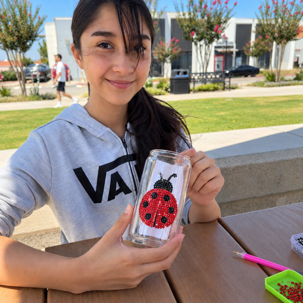A girl holds a clear glass jar decorated with a red and black ladybug design made of small rhinestones.  she decorating bedazzled glass jars with colorful rhinestones