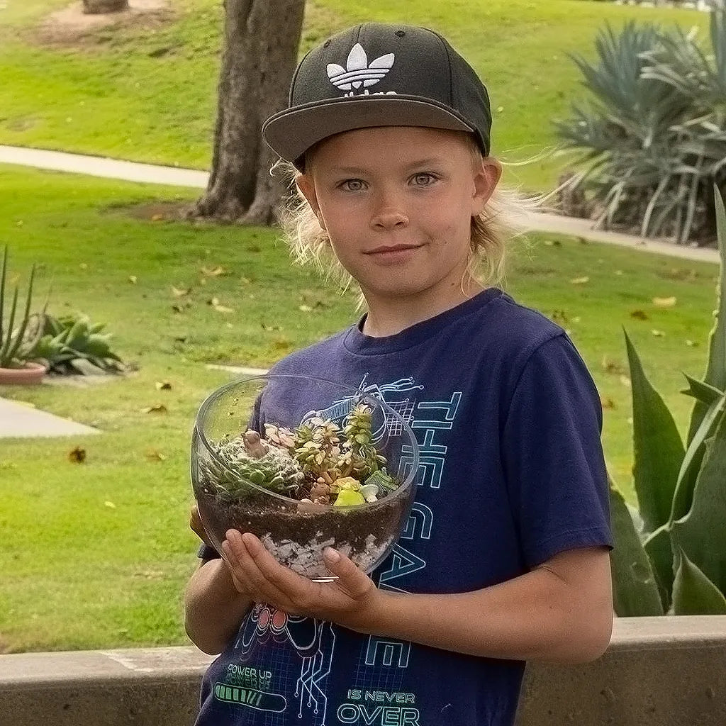 Child proudly holding a finished succulent terrarium after a creative gardening workshop outdoors.